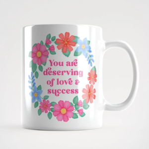Motivational mug with quote: You are deserving of love and success. Pink text with blue, pink and orange floral wreath with green leaves and foliage.